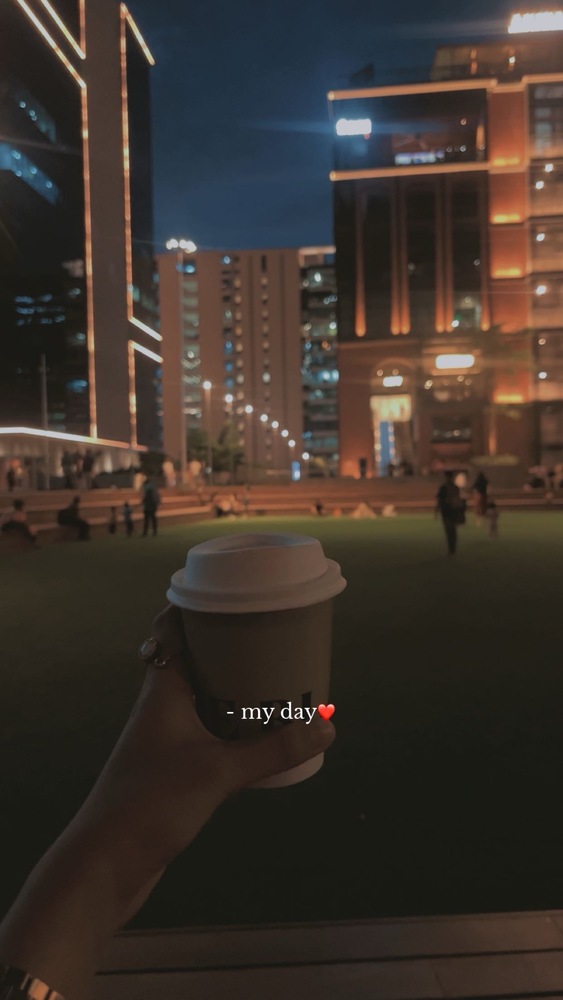 snap showing a hand holding a to-do coffee cup. a green field is in the background surrounded by buildings and lots of ambient lighting